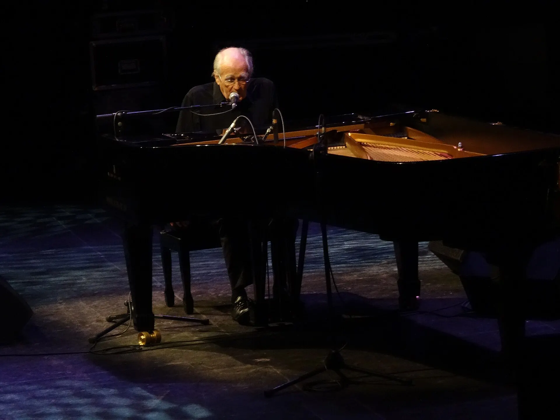 Michel Legrand au piano à queue, chantant au micro, scène plongée dans le noir
